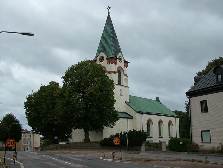 Ödeshögs kyrka med grönt tak, vit fasad och träd runtom.