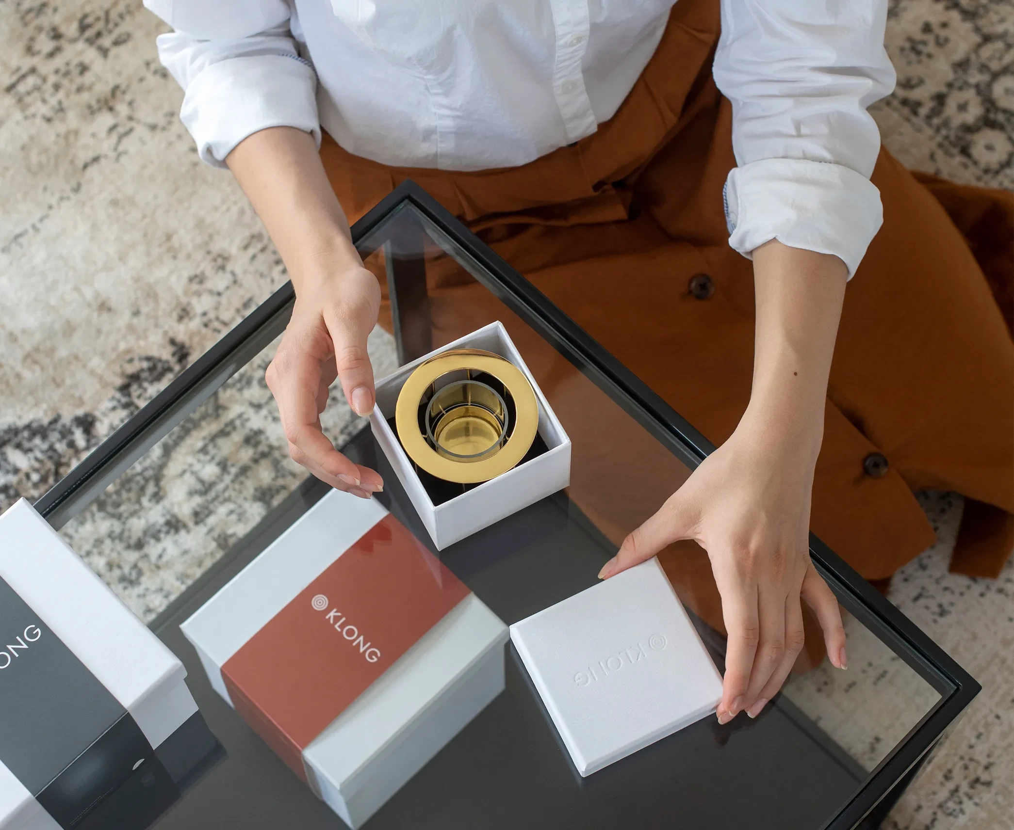 Person unboxing a gold candle holder from a white KLONG-branded gift box on a glass table, showcasing premium packaging design.
