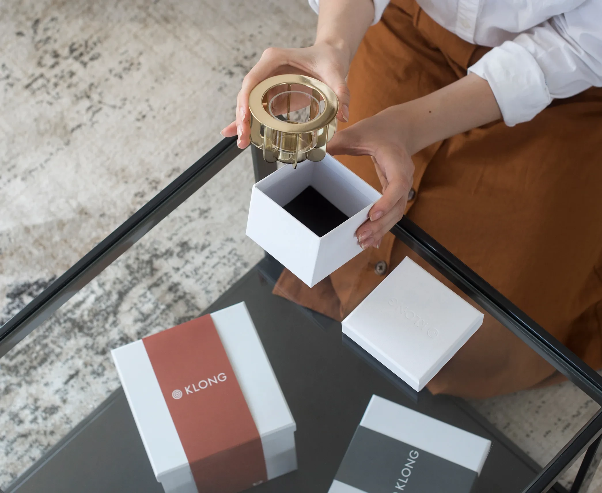 Person holding a gold candle holder above an open white KLONG-branded gift box on a glass table, highlighting elegant product packaging.