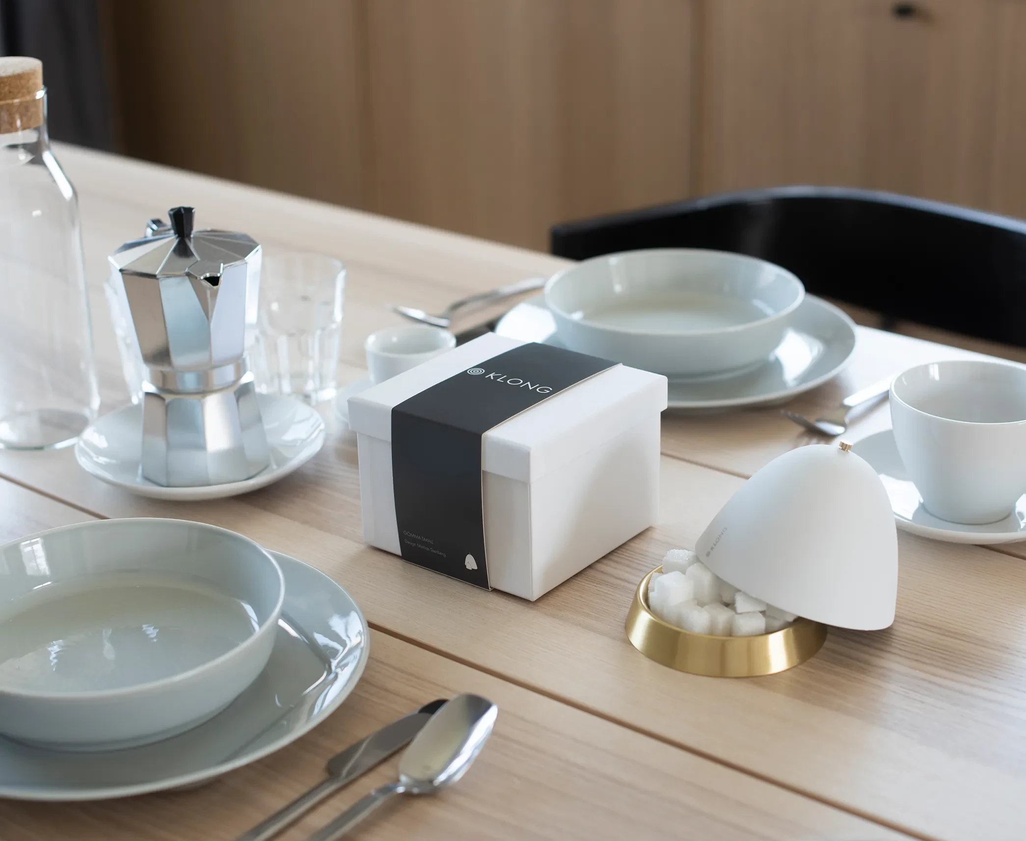 KLONG-branded white gift box with black sleeve placed on a neatly set dining table with coffee maker, cups, plates, and sugar bowl.