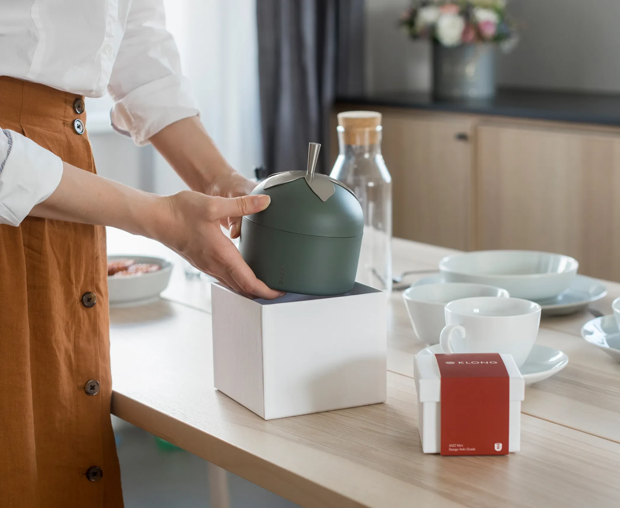 Person unboxing a green apple-shaped KLONG container from a white gift box on a dining table set with cups, plates, and glassware.