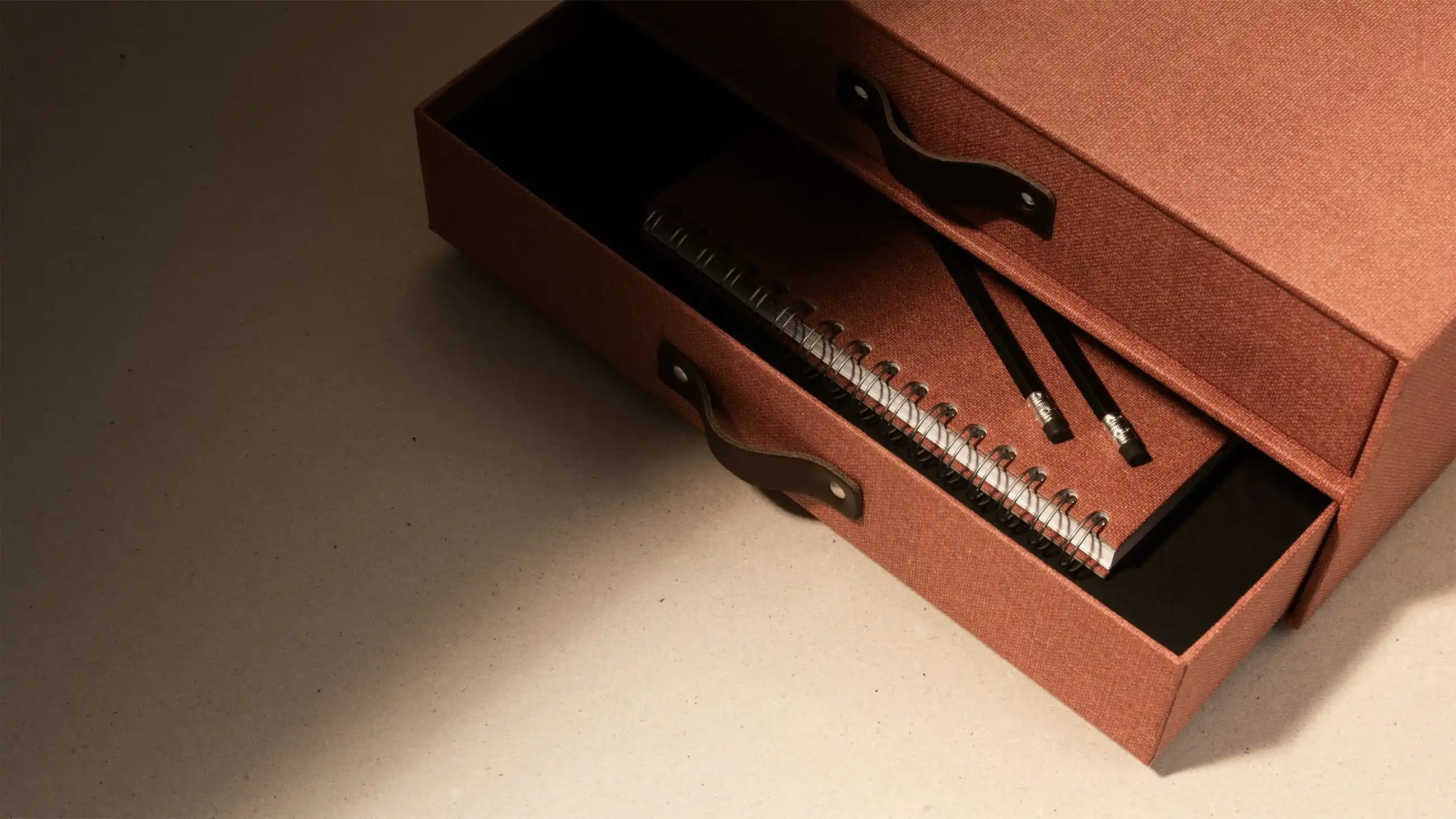 Stylish brown Bigso storage box with leather handles, open drawers revealing a spiral notebook and pencils on a beige surface.