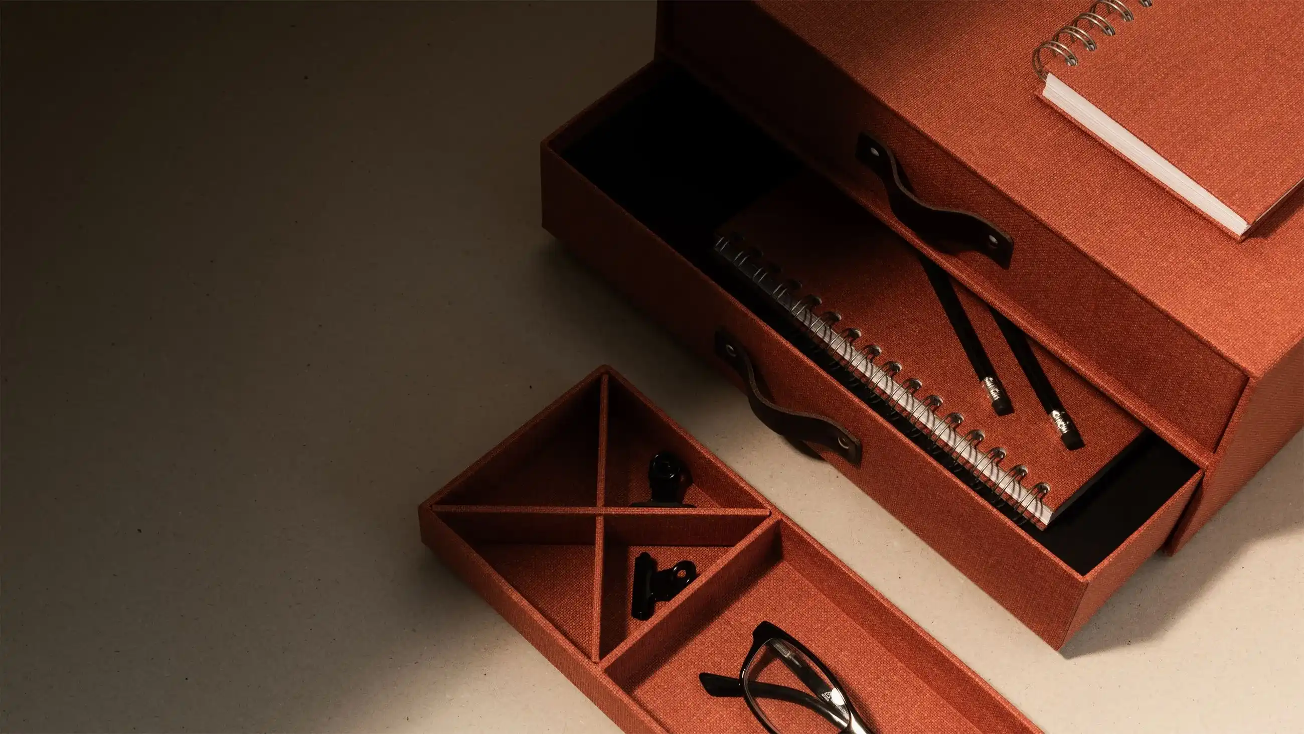 Elegant brown Bigso storage drawers with leather handles, holding notebooks, pencils, eyeglasses, and office clips on a beige desk surface.