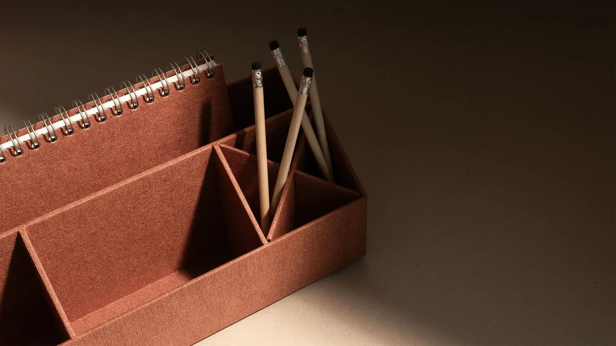 Brown fabric Bigso desk organizer with compartments holding pencils and a spiral notebook, placed on a neutral beige surface.