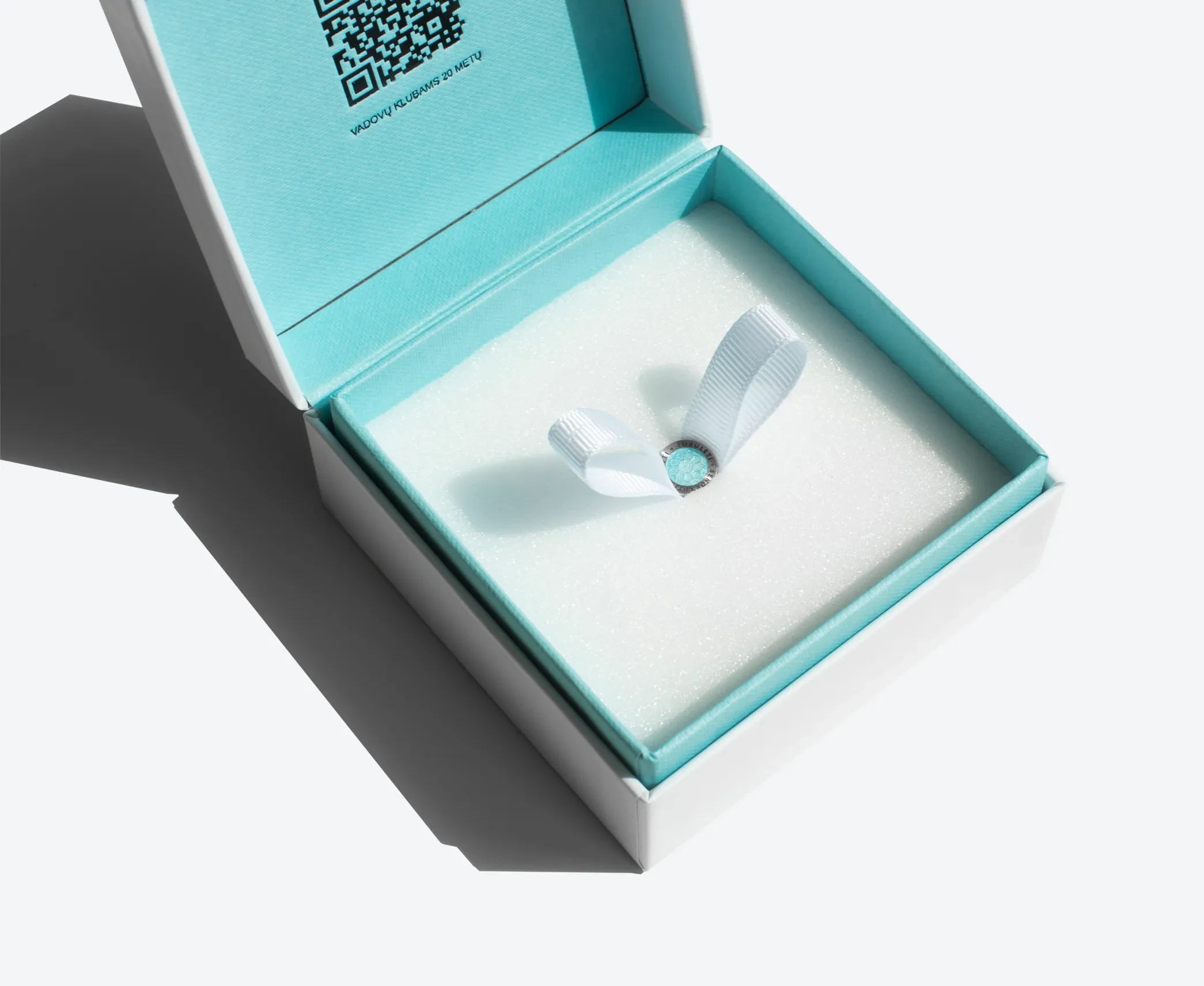 Open hinged lid jewelry box with turquoise interior and foam insert holding a ring with a white ribbon loop.