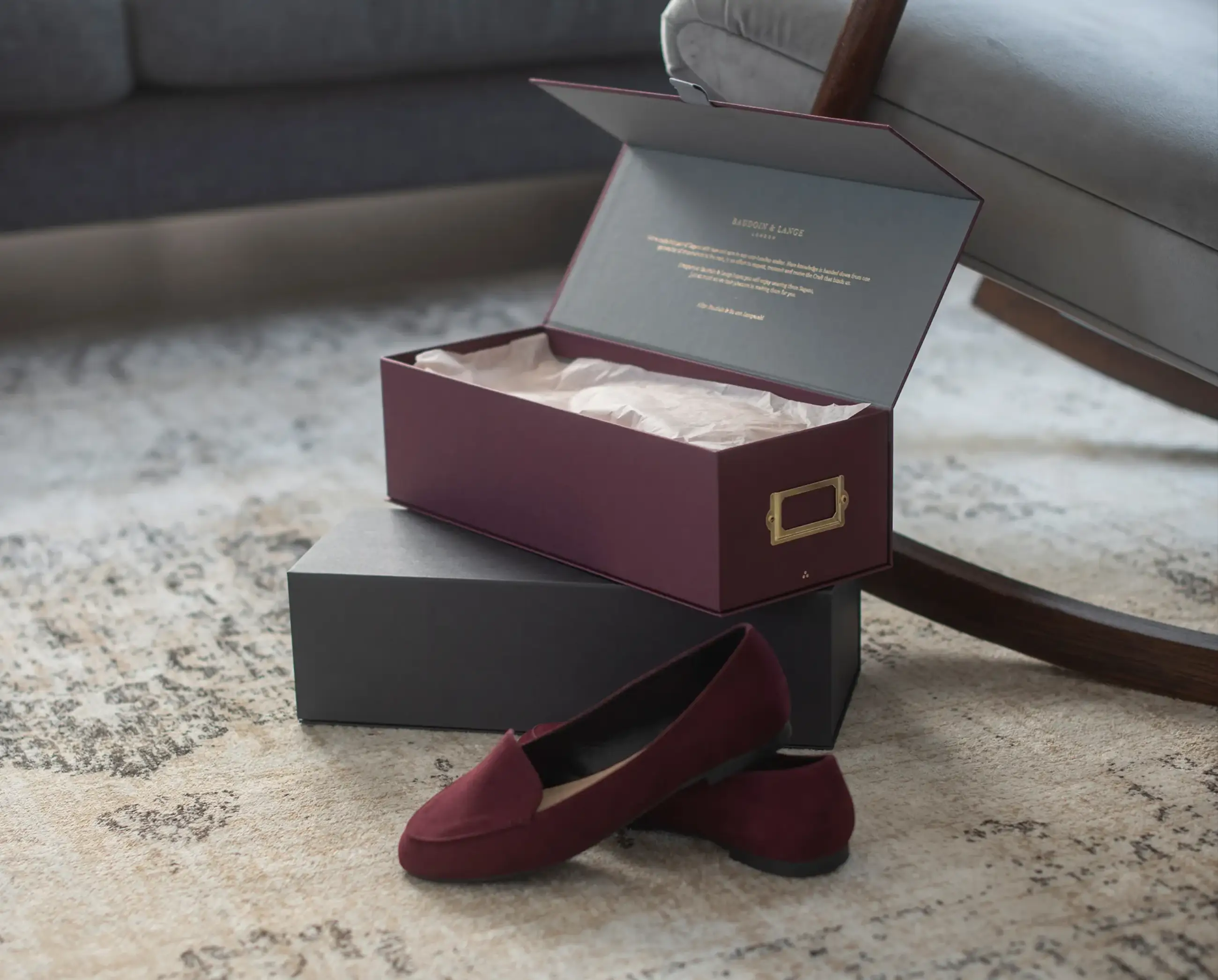 A burgundy magnetic box with a hinged lid, open to display tissue-wrapped shoes, placed on a carpet beside another box and a pair of shoes.