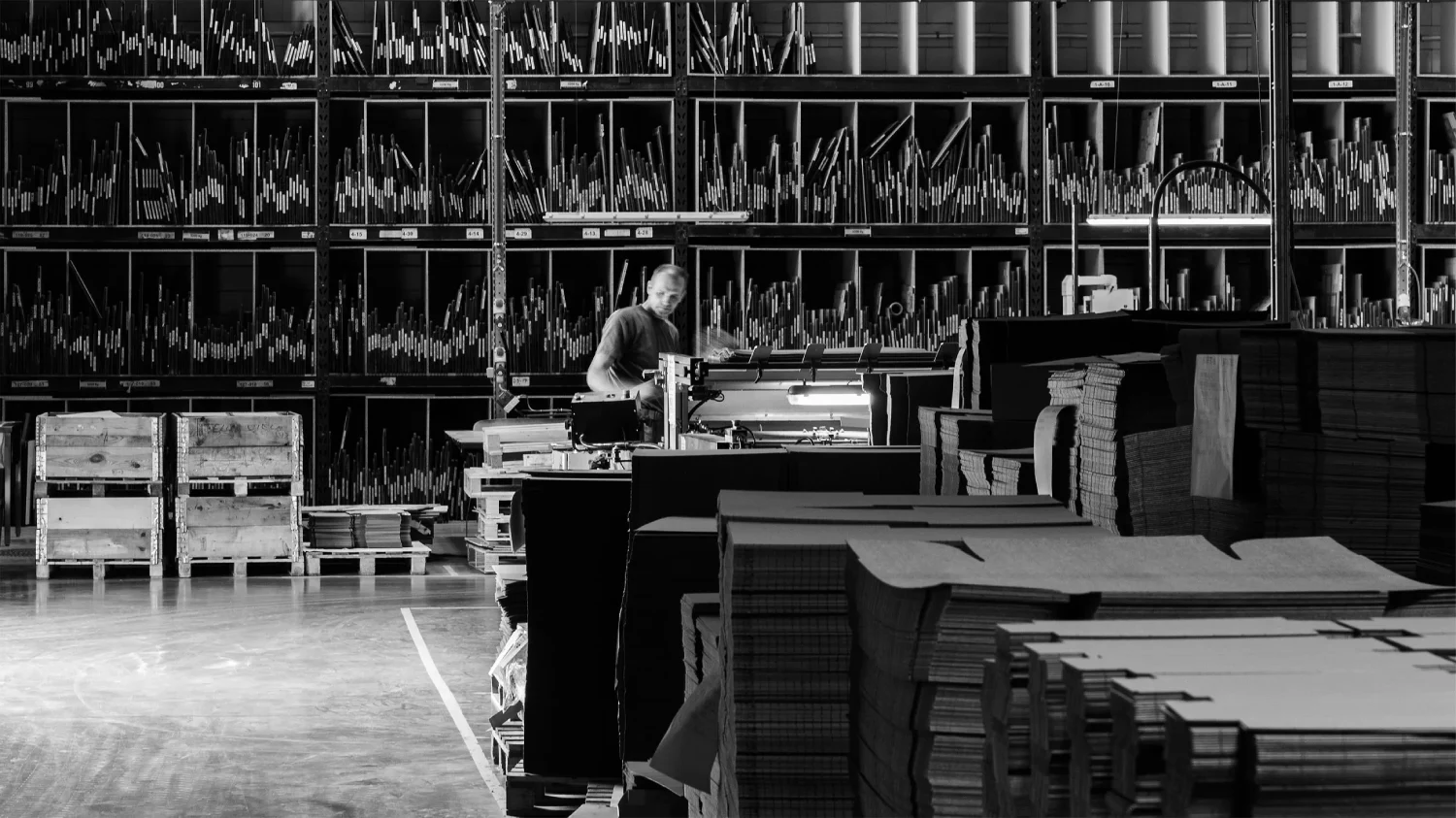 Wide view of Bigso’s factory floor with shelves of tools, stacks of recycled cardboard, and a worker operating precision machinery. Showcasing sustainable packaging production, efficient manufacturing, and eco-friendly material handling.