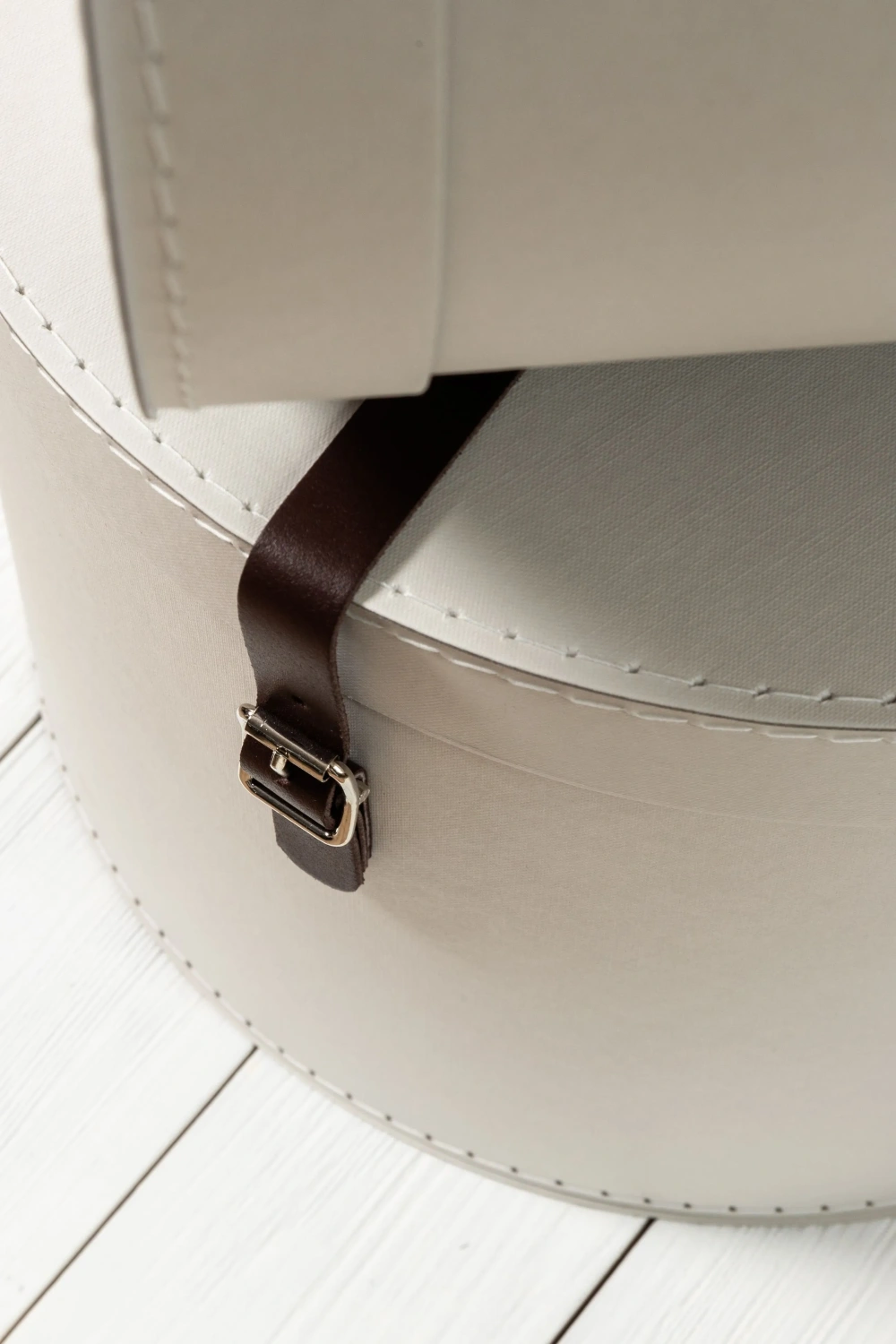 Close-up of a round storage box from Bigso, crafted with cream-colored recycled fiberboard and stitched detailing, featuring a dark brown faux-leather strap with metal buckle. The design highlights Bigso’s classic Scandinavian craftsmanship, blending sustainable materials with timeless style for elegant home and office organization.