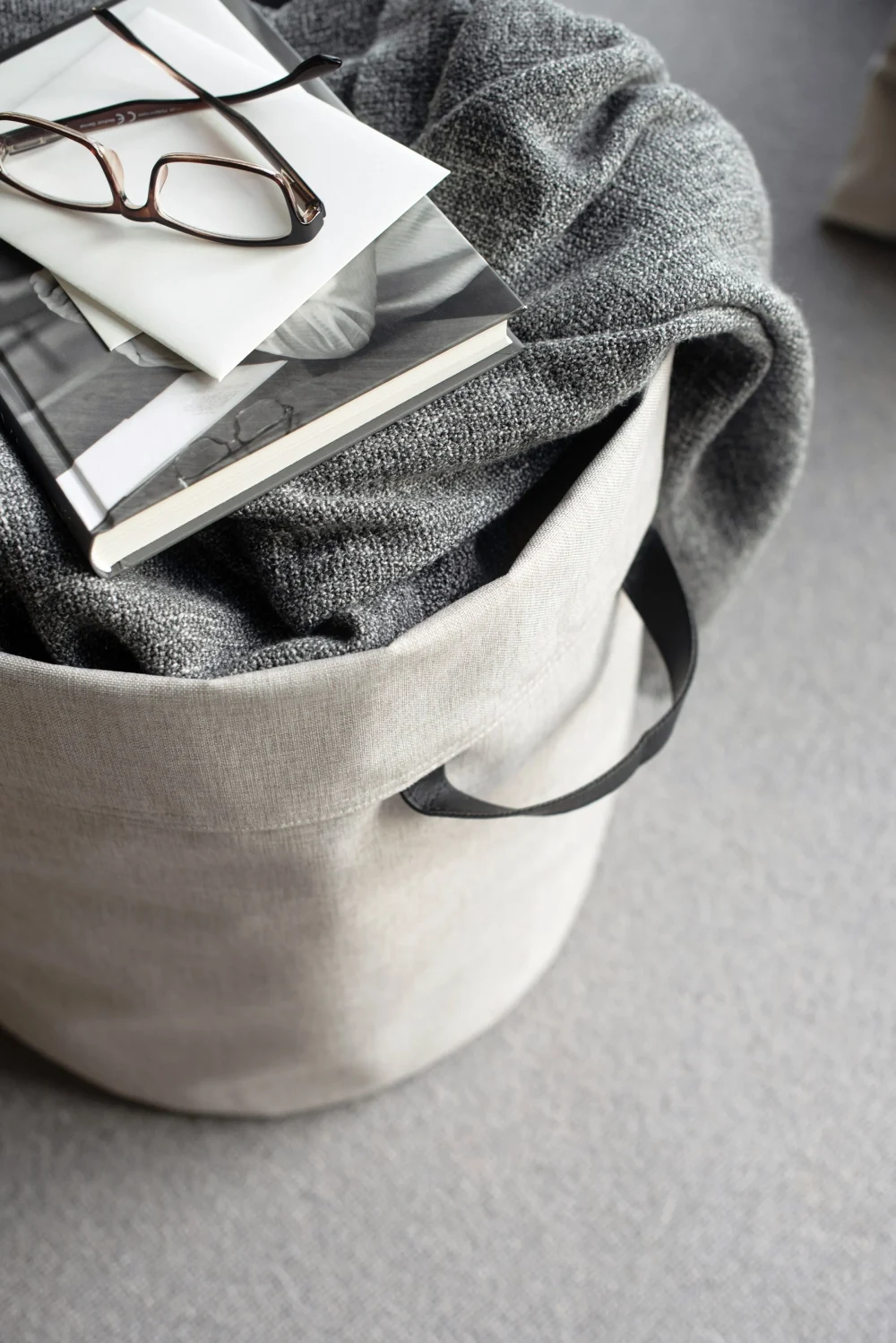 Soft beige fabric storage basket from Bigso, filled with a cozy grey knitted blanket and topped with a book, envelope, and reading glasses. This eco-friendly soft storage solution with sturdy black handles offers stylish and practical organization for living rooms, bedrooms, or offices.