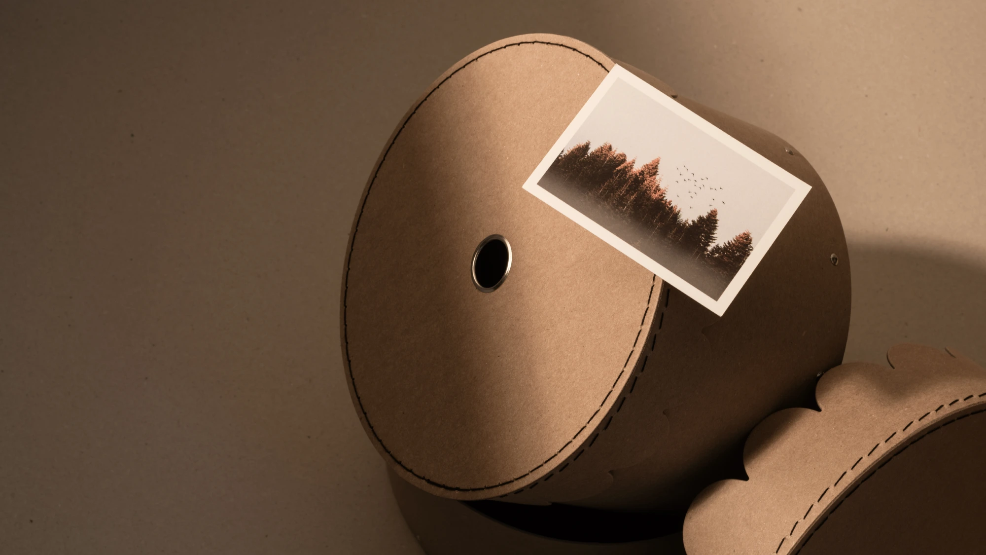 Eco-friendly round storage box made from recycled cardboard with visible stitched edges, photographed with a postcard resting on top. Minimalist sustainable packaging design combining durability, functionality, and aesthetic appeal.