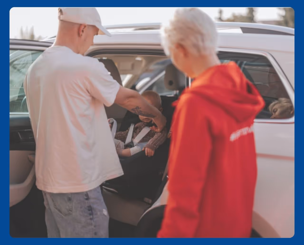 An adult securing a child into a car seat while another adult stands nearby.