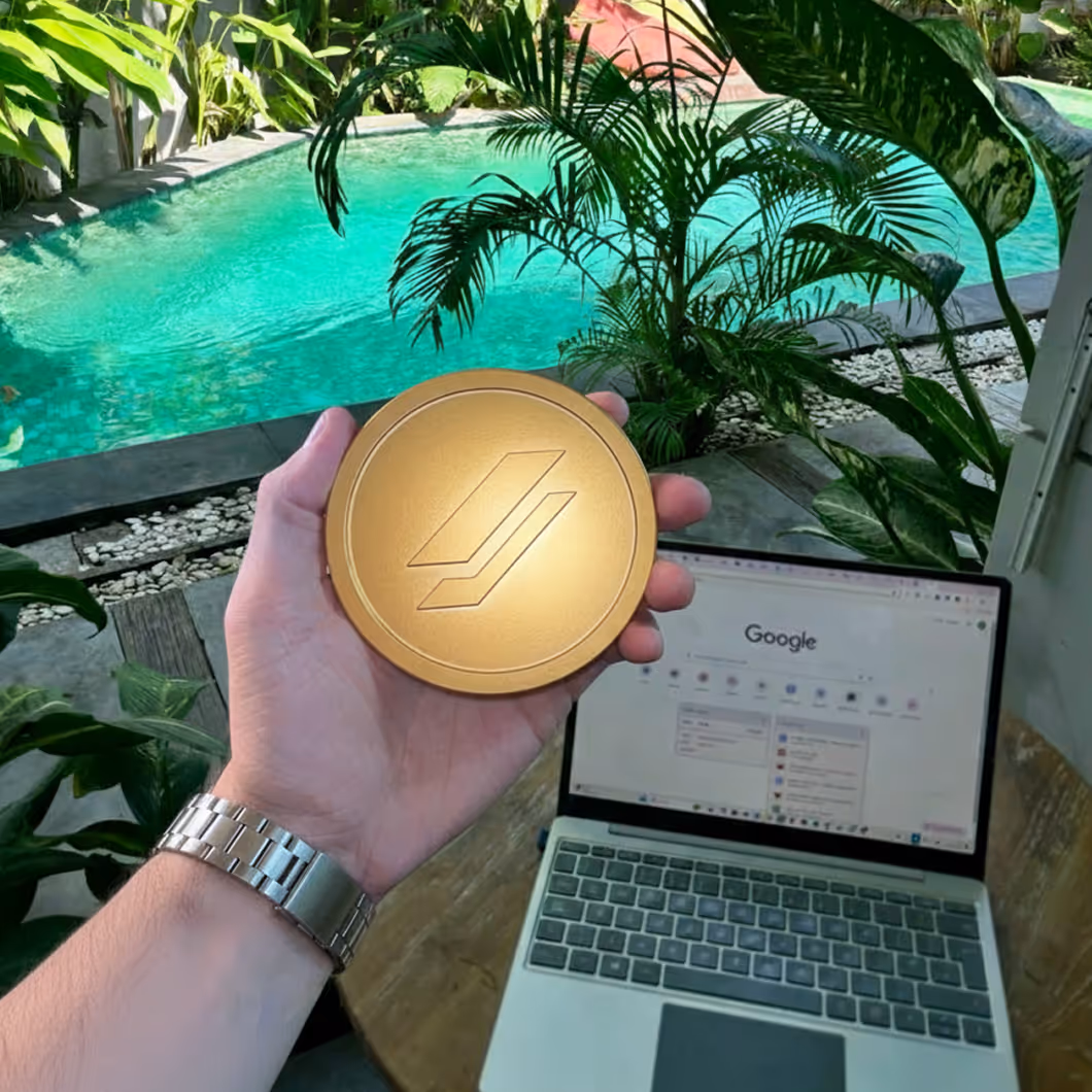 Hand holding a round gold object with a geometric logo in front of a laptop and a turquoise pool surrounded by plants.