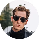 Man wearing black sunglasses and a black jacket with a blurred green outdoor background.