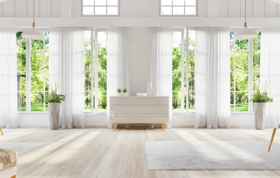 Bright living room with large white-framed windows, sheer curtains, and view of green trees outside