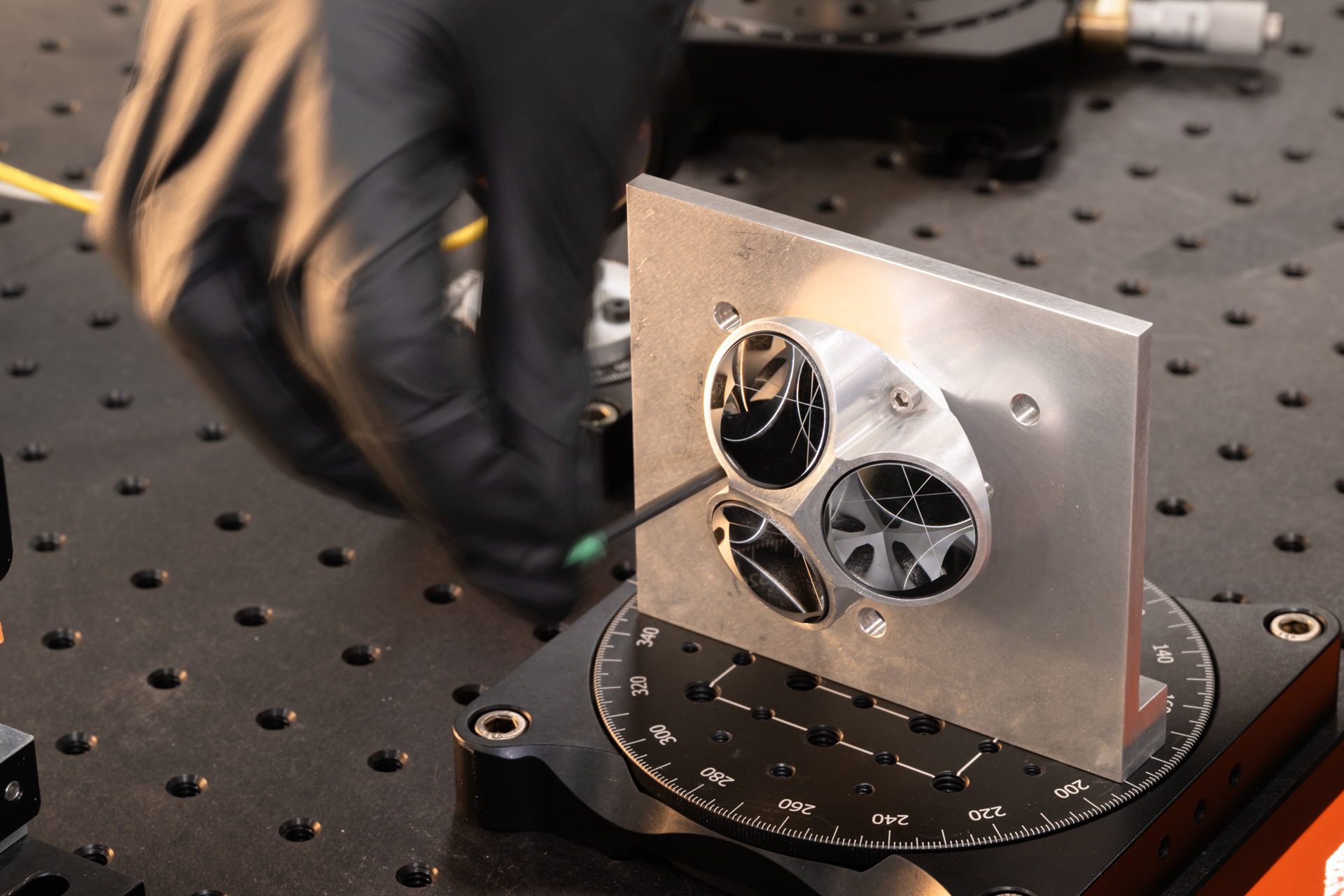 Silver laser retroreflector array is on a table as a black-gloved hand tinkers with the top corner.