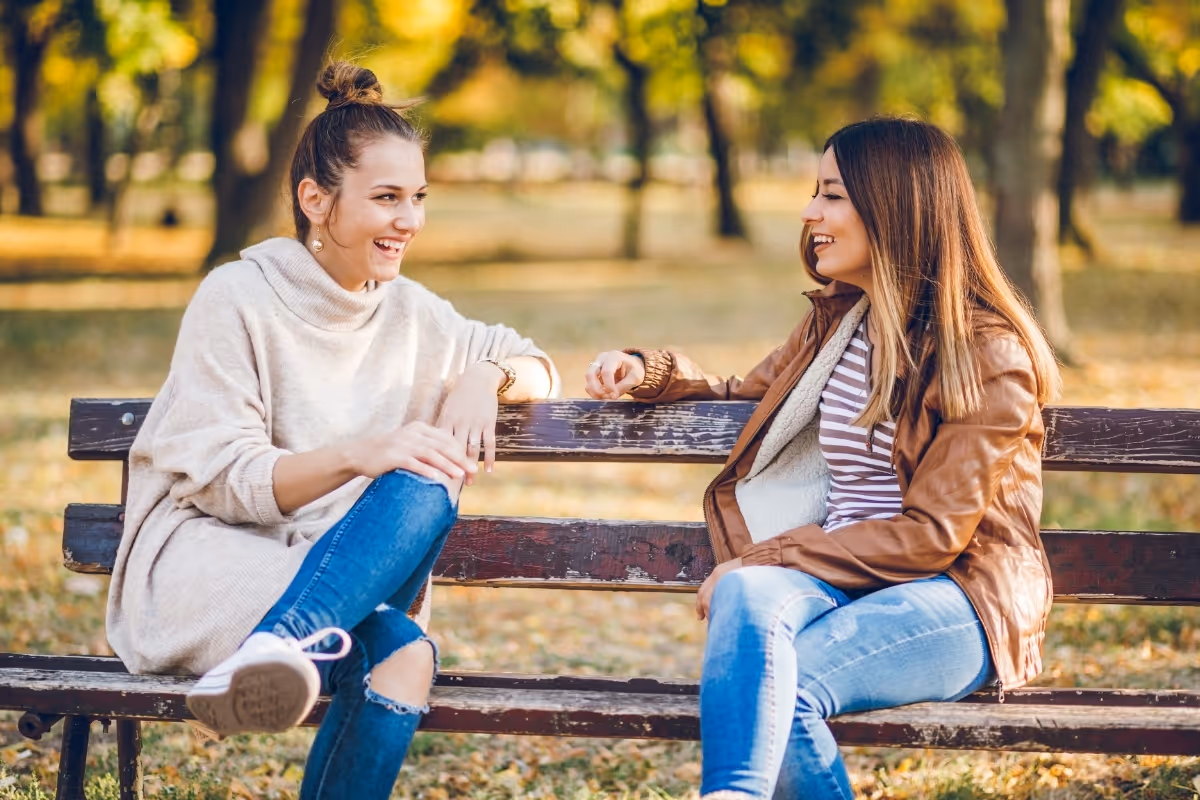 two young women on a bench in a park smiling and talking