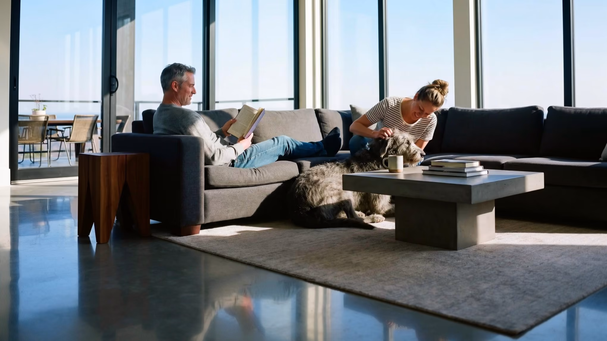Man sitting on a gray couch reading a book while a woman leans over the couch petting a large gray dog lying on a rug by a modern coffee table with books and a cup, in a bright room with large windows.
