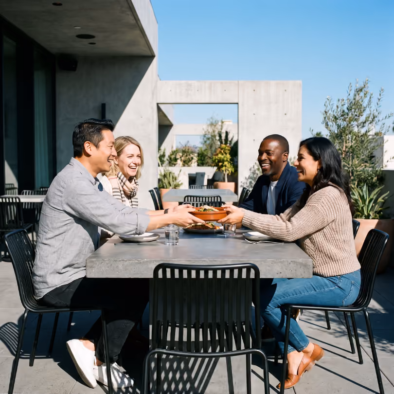 Two couples laughing and passing a dish while sitting outdoors at a modern concrete table with black chairs under a clear blue sky.
