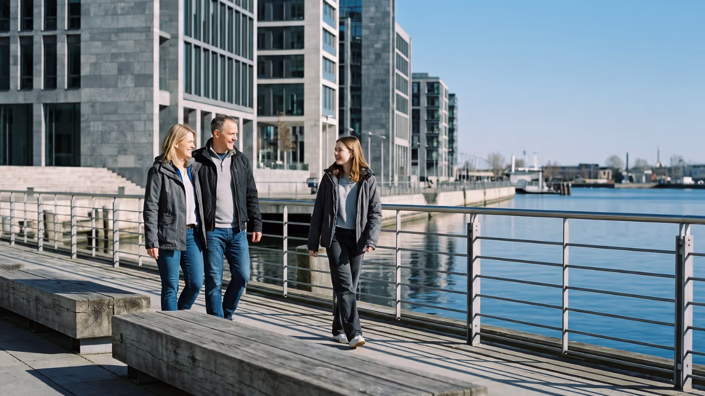 A family of three, two adults and one teenager, walking along a waterfront promenade beside modern buildings under a clear blue sky.