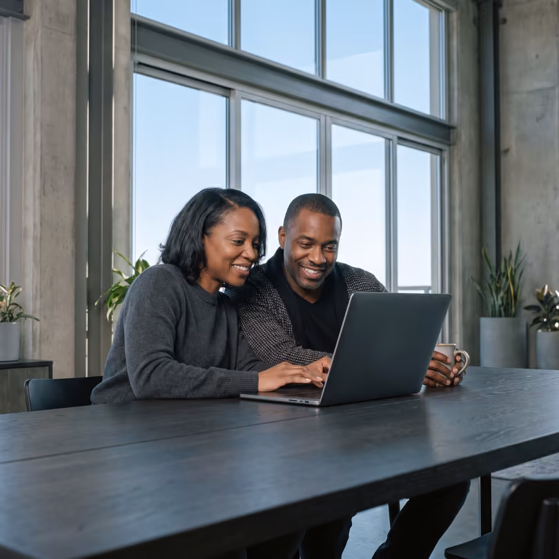 Smiling couple sitting at a large table using a laptop together in a modern room with large windows and plants.