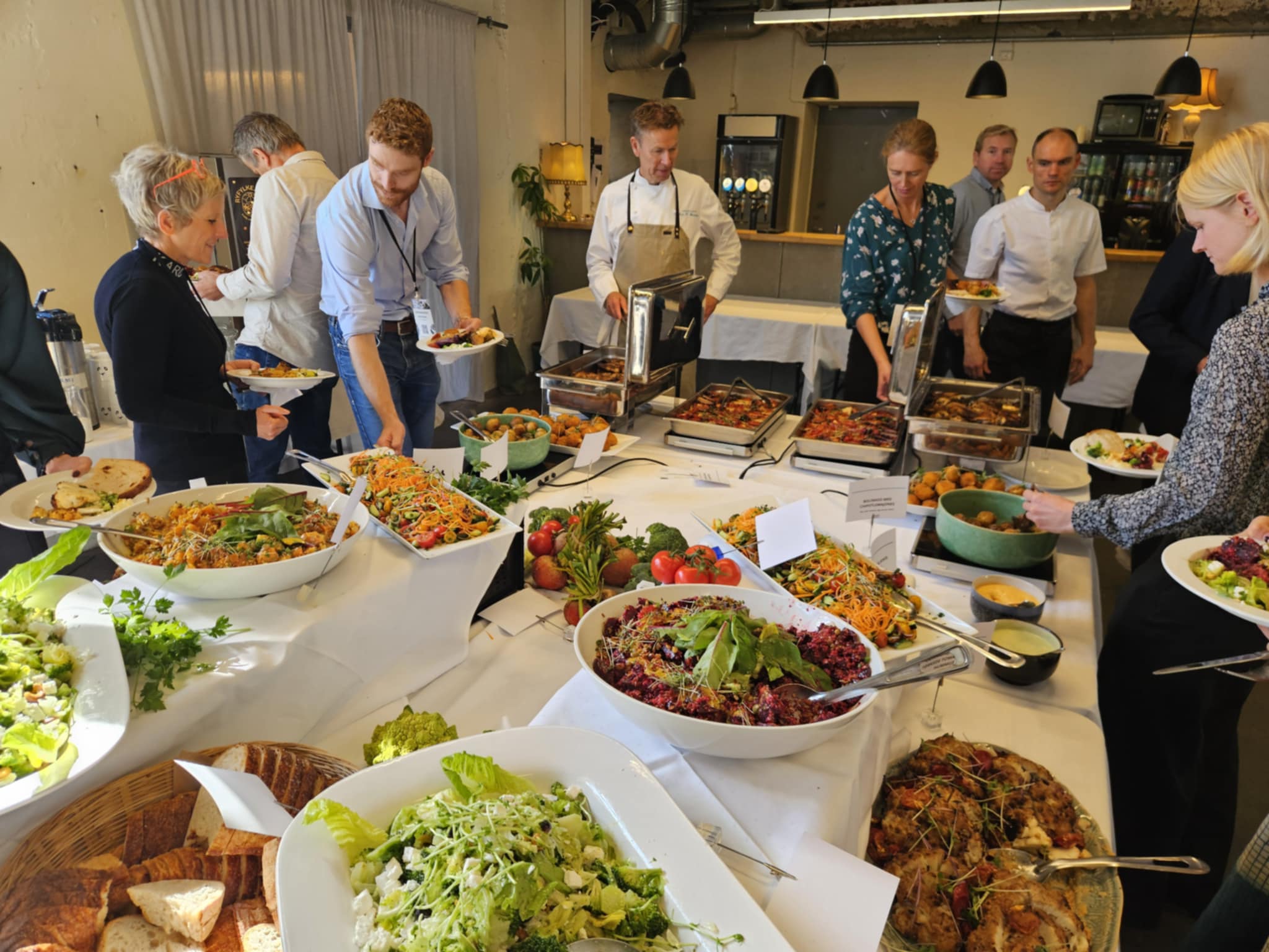 Folk som tar mat fra et buffetbord med salater, grønnsaker, brød og varme retter i en lys kantine.