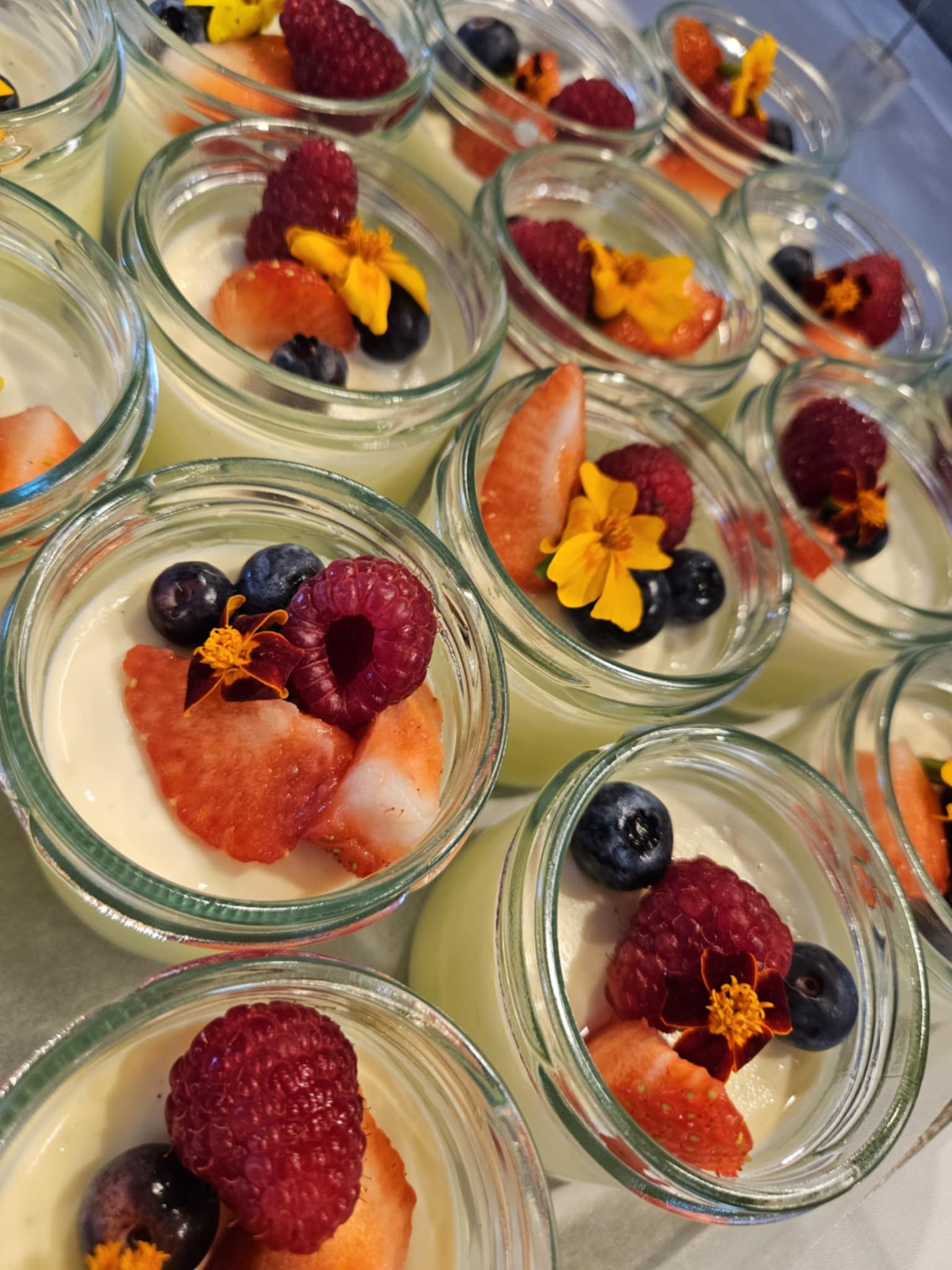 Små glasskrukker med vaniljepudding toppet med bringebær, blåbær, jordbær og spiselige blomster.