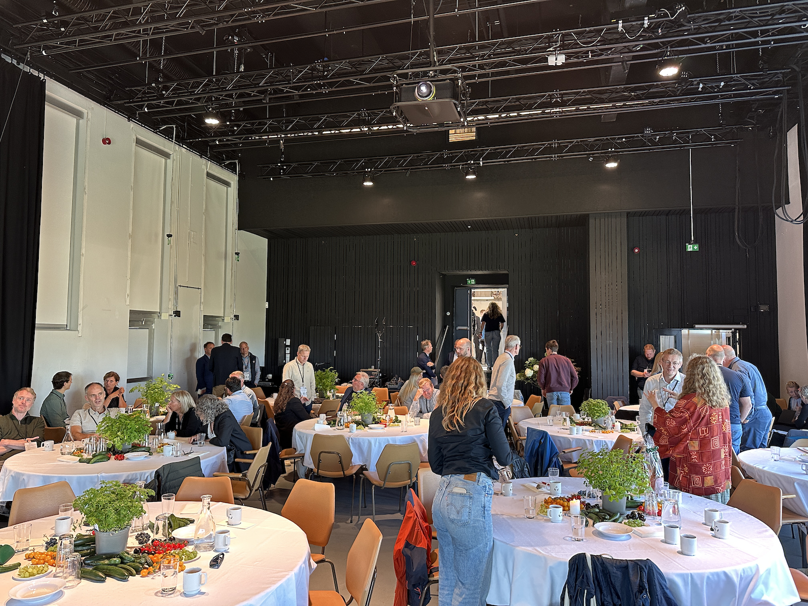 Folk i et konferanserom med runde bord dekket med hvite duker, planter og drikkevarer, sosialiserer og snakker.