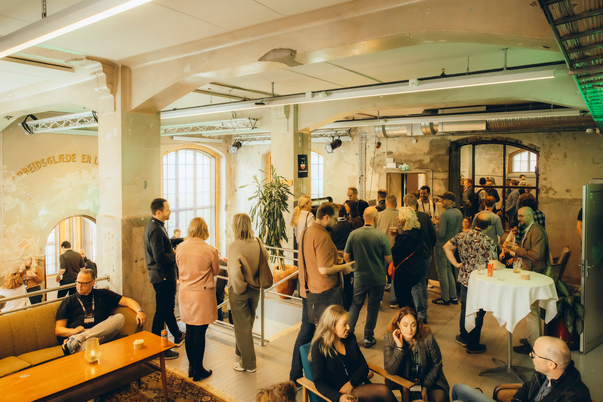 Mennesker i samtale under et sosialt arrangement i et lyst, moderne lokale med store vinduer og industrielt preg.