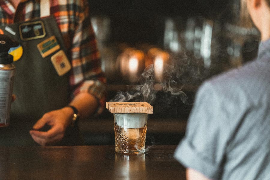An Old Fashioned cocktail in a rocks glass with a charred cedar wood smoker top resting on the rim, releasing wisps of smoke into the amber liquid.