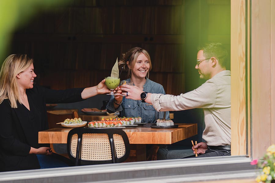 Two women and a man cheers their cocktails over a table with plated sushi rolls. The group sits next to an open window leading out to the patio.