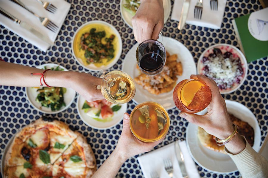 Four hand cheers their wine and cocktail glasses of a table set with happy hour food items.