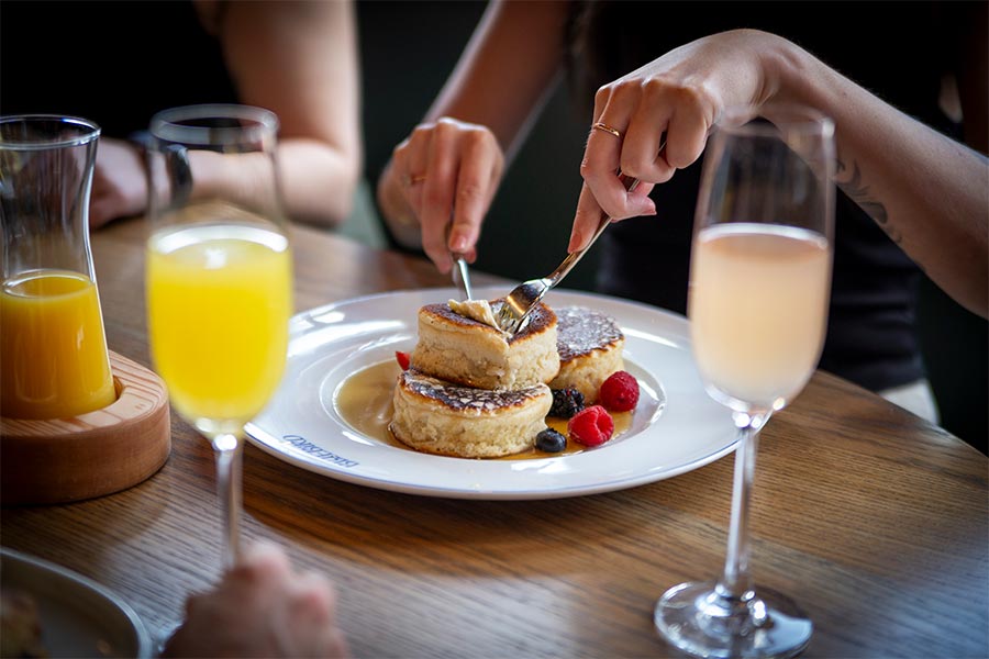 A fork and knife cutting into a stack of thick fluffy pancakes covered in maple syrup with a few berries on the side.