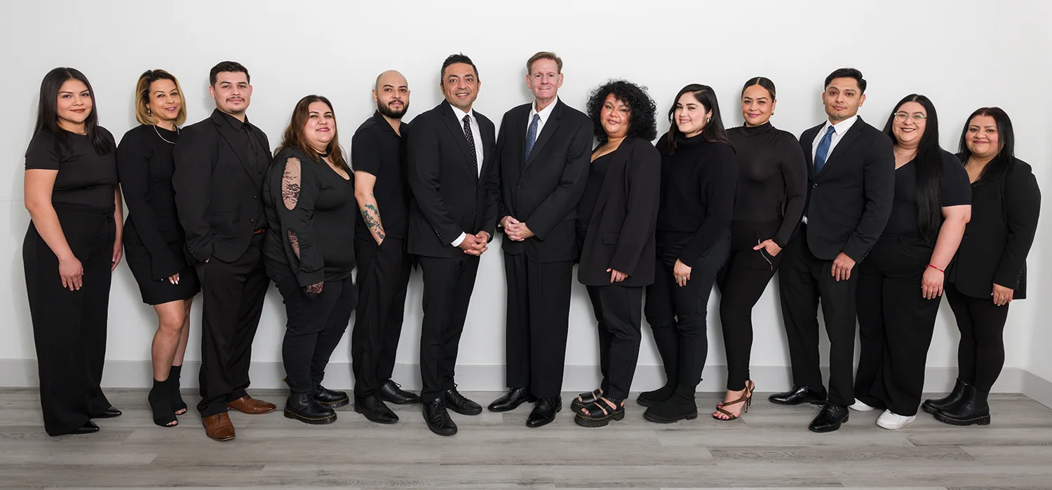 Group of fourteen professionally dressed staff at California Attorney Group standing in a row against a white wall.