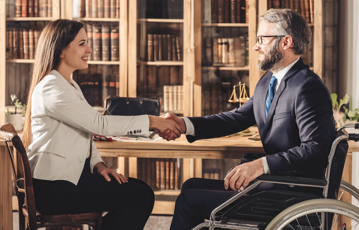 Un abogado de lesiones personales en traje y silla de ruedas le da la mano a una clienta dentro de una oficina legal, simbolizando la confianza, la defensa y la accesibilidad.