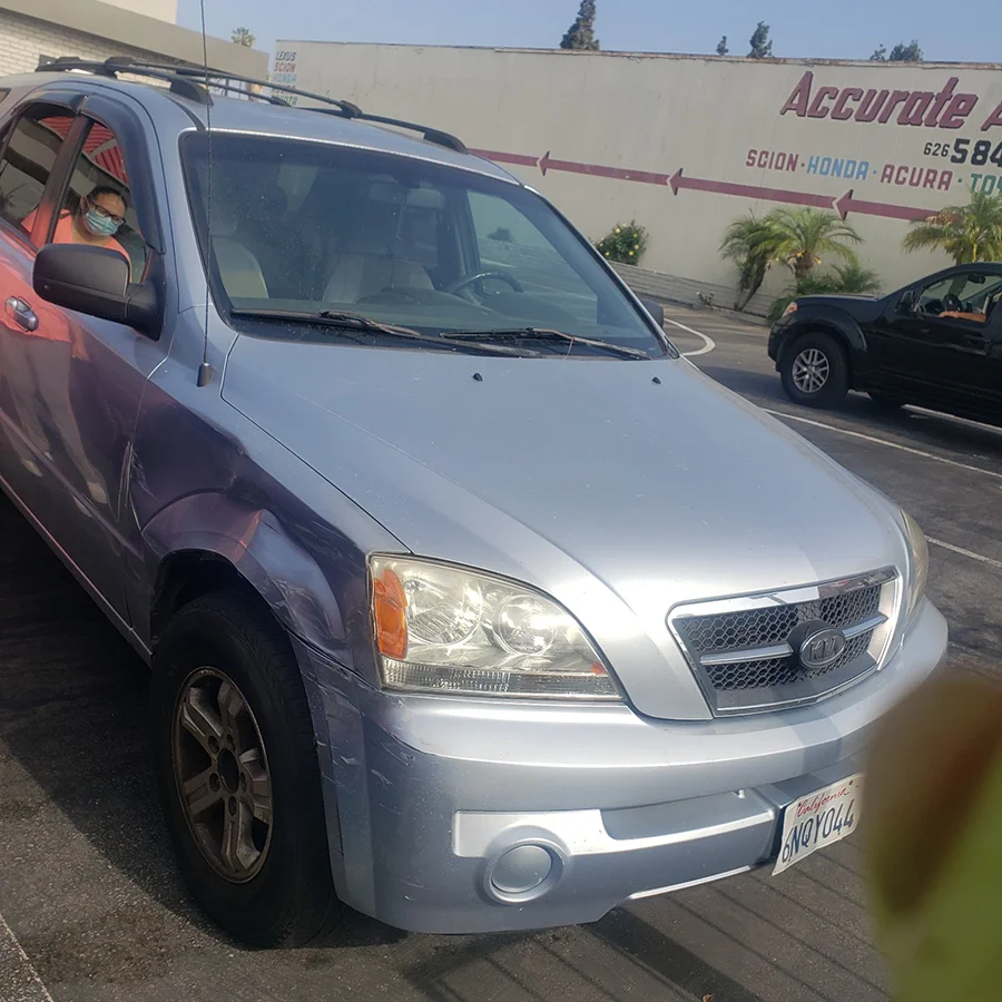 A silver SUV with visible front and side damage is parked in a lot outside an auto repair shop. The driver’s side fender shows dents and scratches from a collision. A person wearing a mask is seated in the vehicle, and other cars and a building marked “Accurate Auto” are seen in the background.