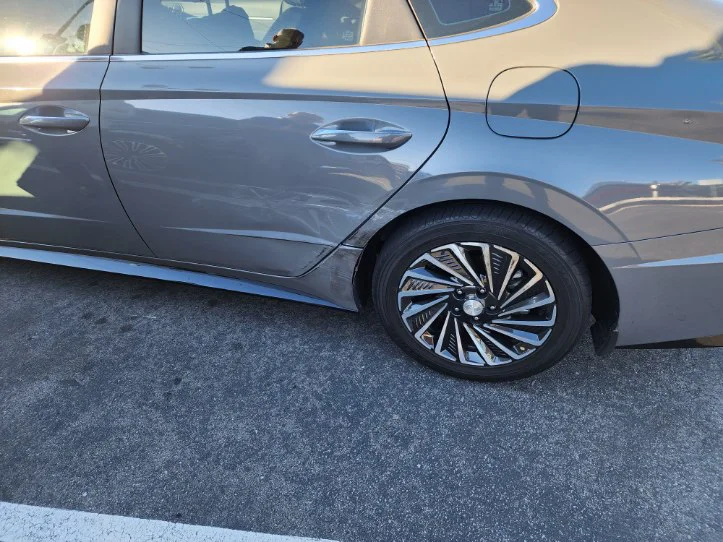 A gray sedan with visible damage on the right rear door and wheel area is parked on a street in daylight. The impact marks and dented panel show the aftermath of a side collision, consistent with a car accident scenario.