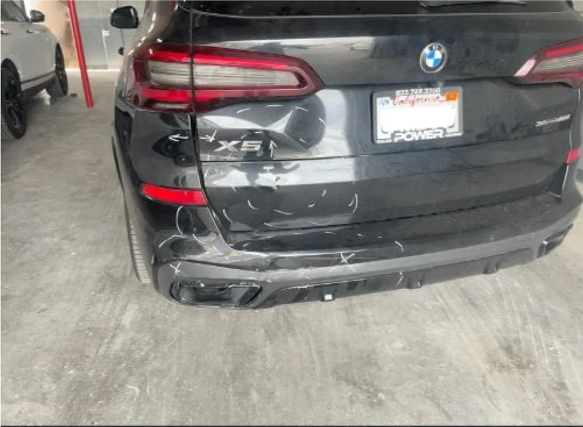 A black BMW X5 SUV shows visible rear-end damage with scratches and impact marks on the bumper and trunk. The vehicle is parked in a garage, illustrating the aftermath of a rear-end collision.
