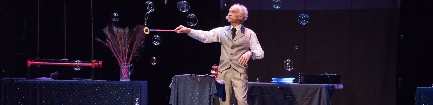 An elderly man in a beige vest and pants blowing soap bubbles on a stage with tables and decorations.