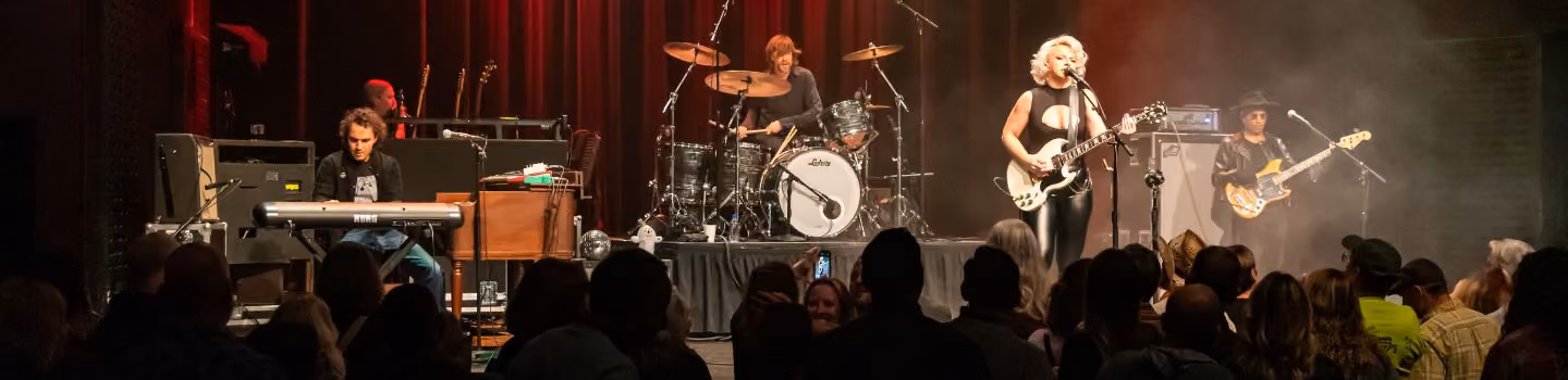 Band performing live on stage with keyboardist, drummer, female guitarist and singer, and bassist in front of an audience.