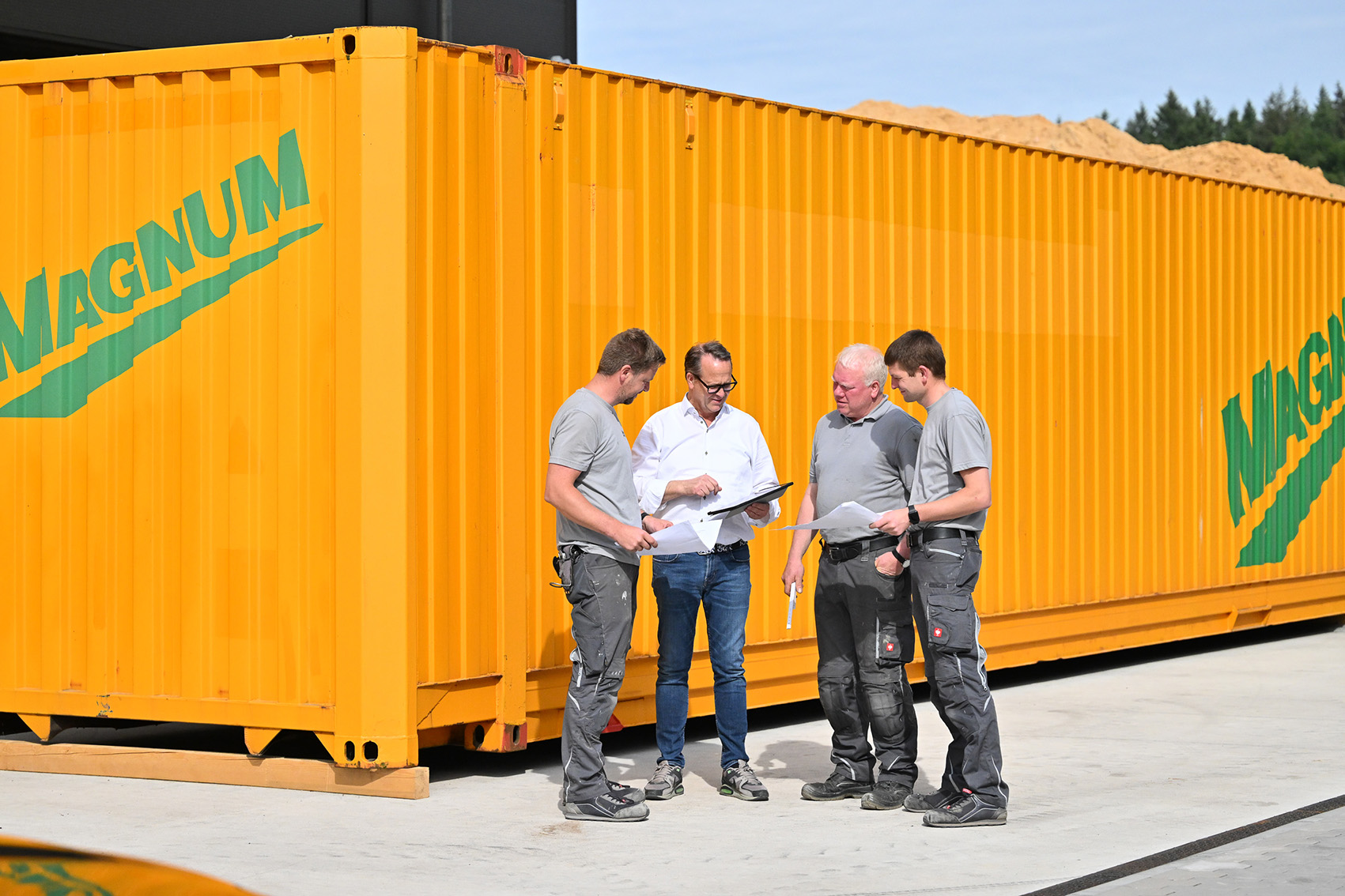 Vier Männer in Arbeitskleidung stehen vor einem großen orangefarbenen Container mit der Aufschrift Magnum und besprechen Unterlagen.