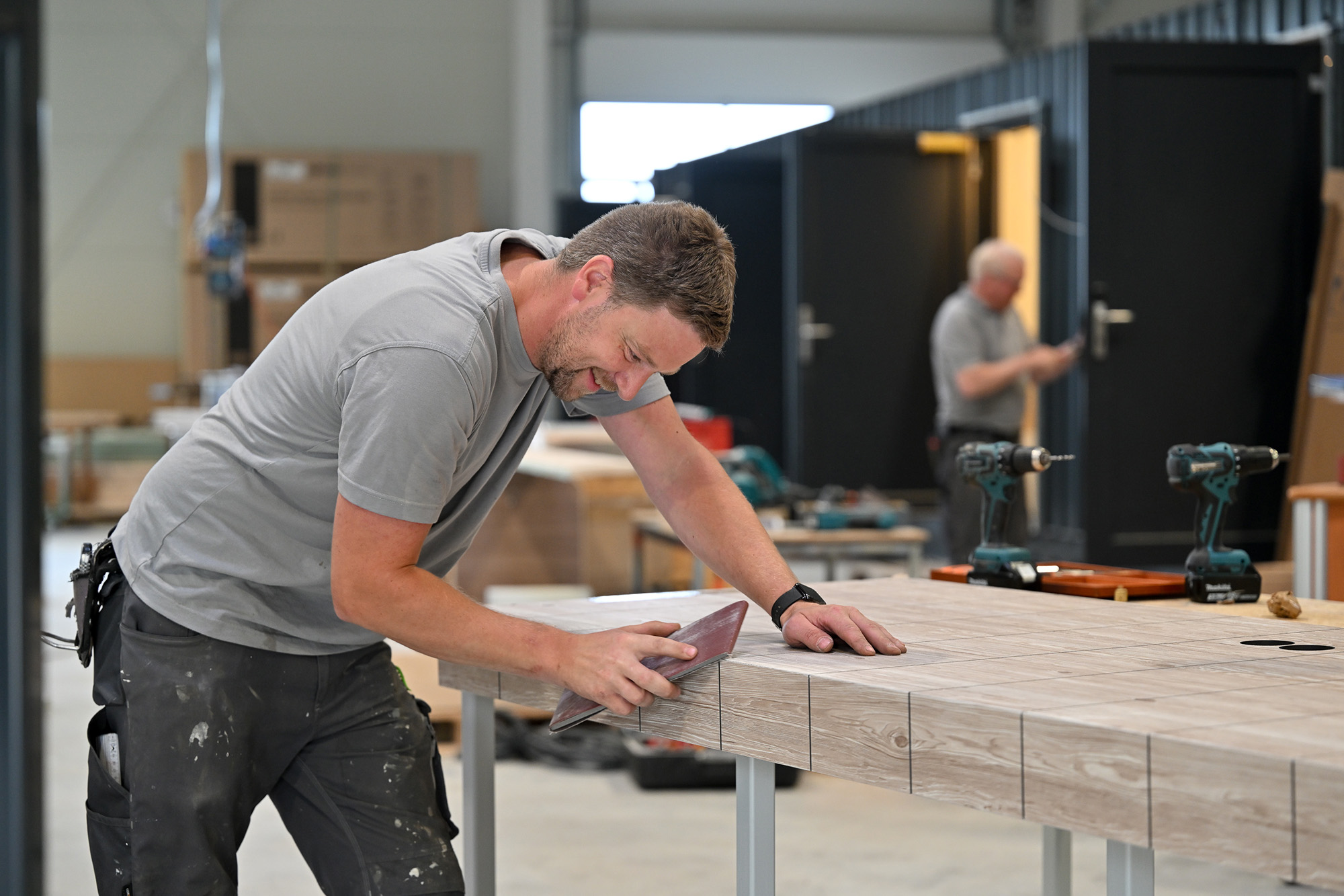 Handwerker schleift eine große Holzplatte in einer Werkstatt, im Hintergrund steht eine weitere Person neben zwei Akkuschraubern.