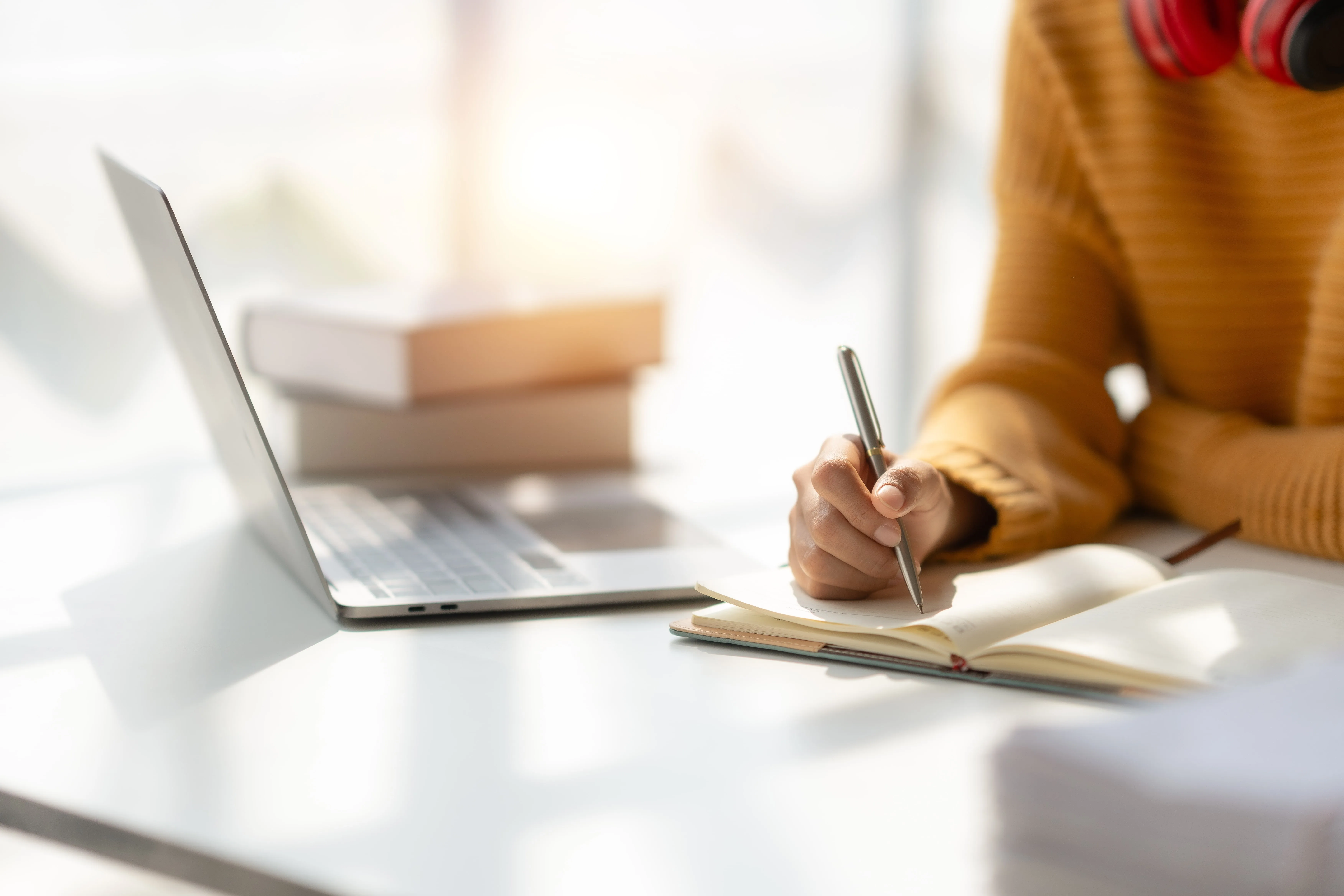 A person is writing in a notebook in front of a laptop