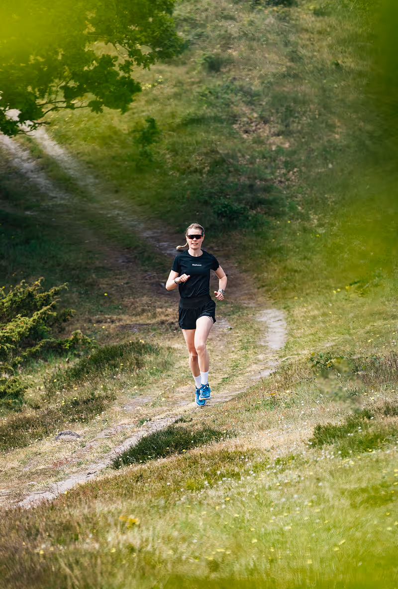 Kvinde løber på en grussti i et grønt, kuperet naturområde iført sort tøj og blå sko.