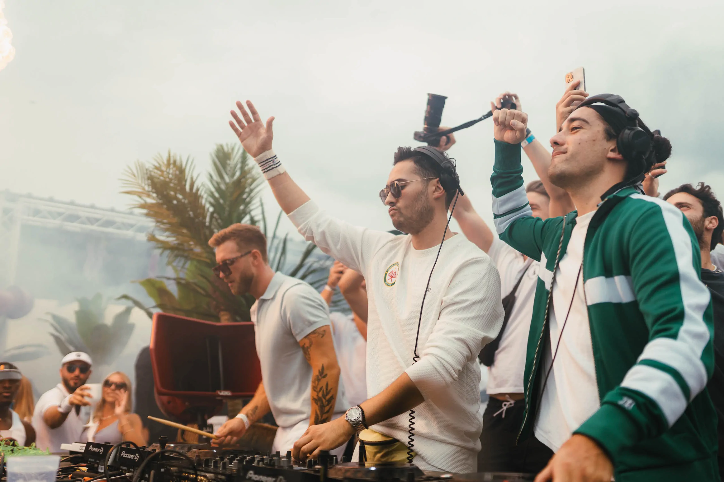 Zwei DJs mit Kopfhörern legen bei einem Outdoor-Event auf, während das Publikum im Hintergrund feiert.