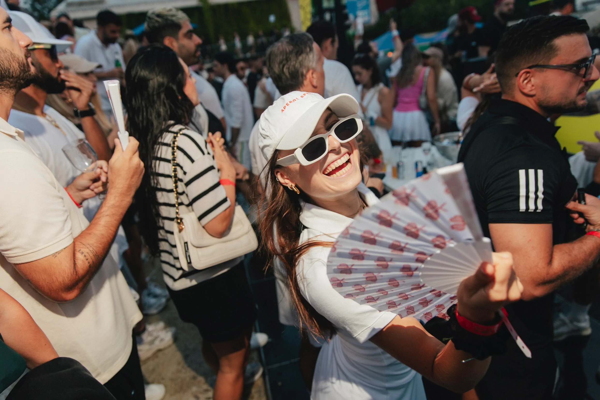 Fröhliche Frau bei einer Menschenmenge hält einen Fächer und trägt Sonnenbrille und weiße Kappe.