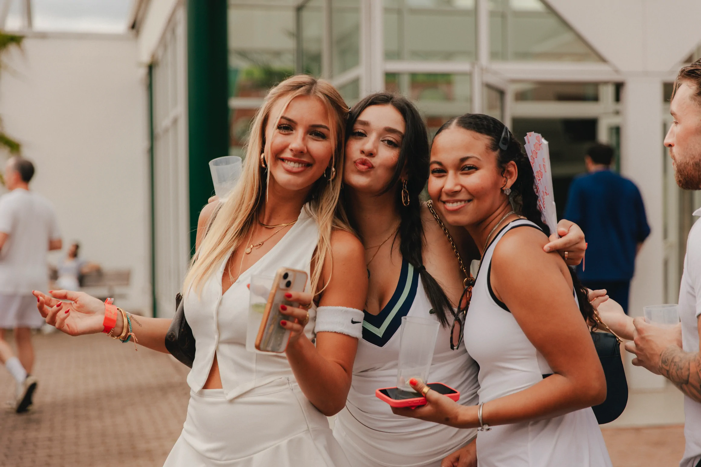 Drei Frauen lachen und posieren zusammen bei einer Veranstaltung im Freien, jede hält ein Getränk und ein Smartphone in der Hand.