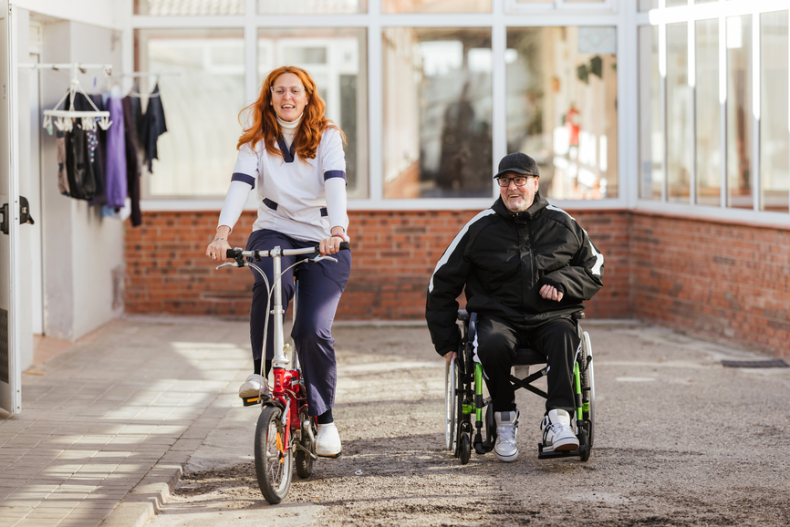 Kvinna på cykel och man i rullstol utomhus i solsken, aktiva tillsammans