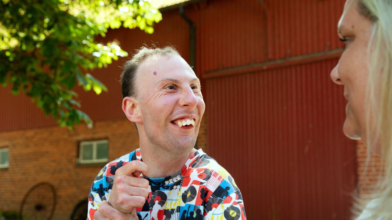 Man i färgglad skjorta skrattar utomhus framför en tegelbyggnad