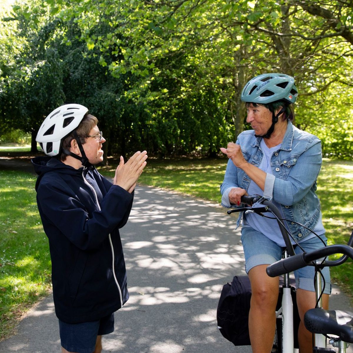 Två personer i cykelhjälmar skrattar tillsammans på en cykelväg i parken