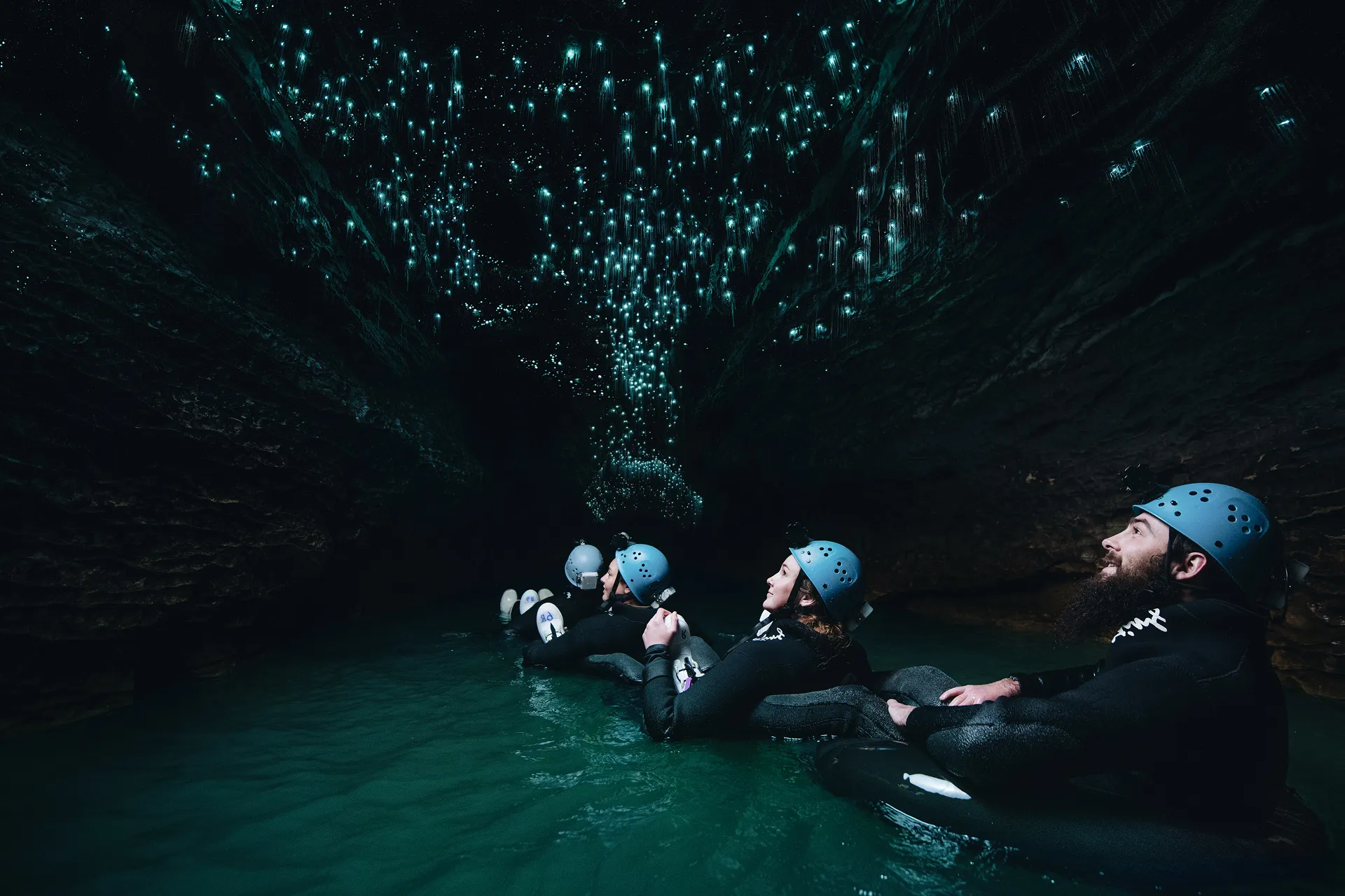 Three people in helmets floating on inner tubes in a dark cave illuminated by glowing blue lights on the ceiling.